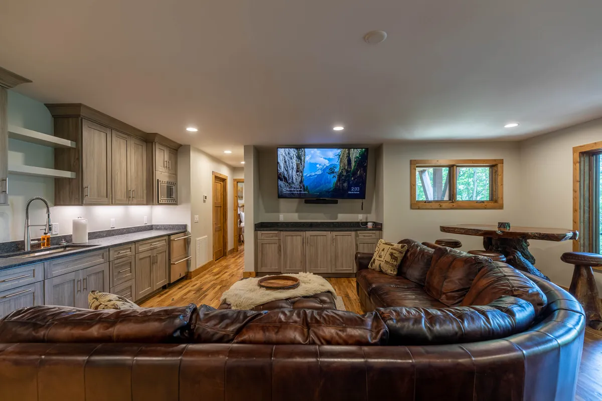 A spacious family room with a large leather sectional sofa, a big-screen TV, a wooden dining table, and kitchen cabinetry visible in the background. The space feels comfortable and welcoming—perfect for movie nights or board games with loved ones.