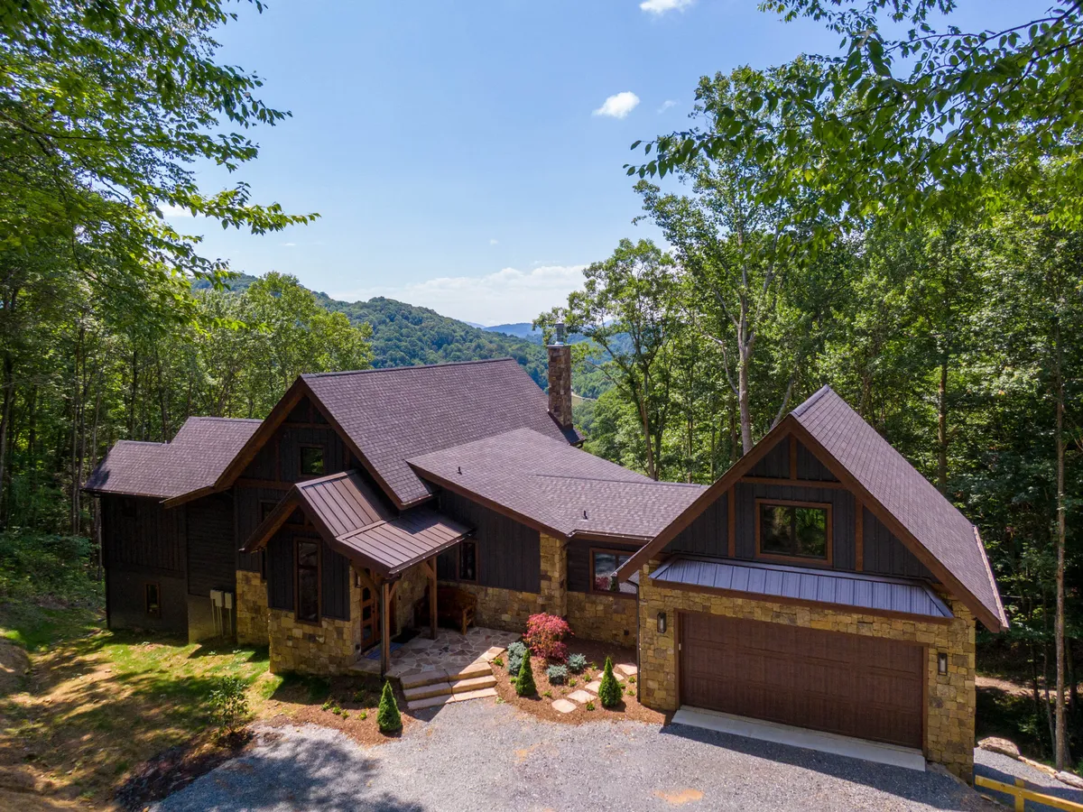 The hero image features a stunning stone facade and sloped roof of a brand new mountain home, nestled amid towering trees. The inviting front porch overlooks a meticulously landscaped path, creating a welcoming entry. The setting is tranquil, with hints of majestic mountain views peeking through the woods, and the atmosphere is perfect for family gatherings, outdoor barbecues, and relaxation.