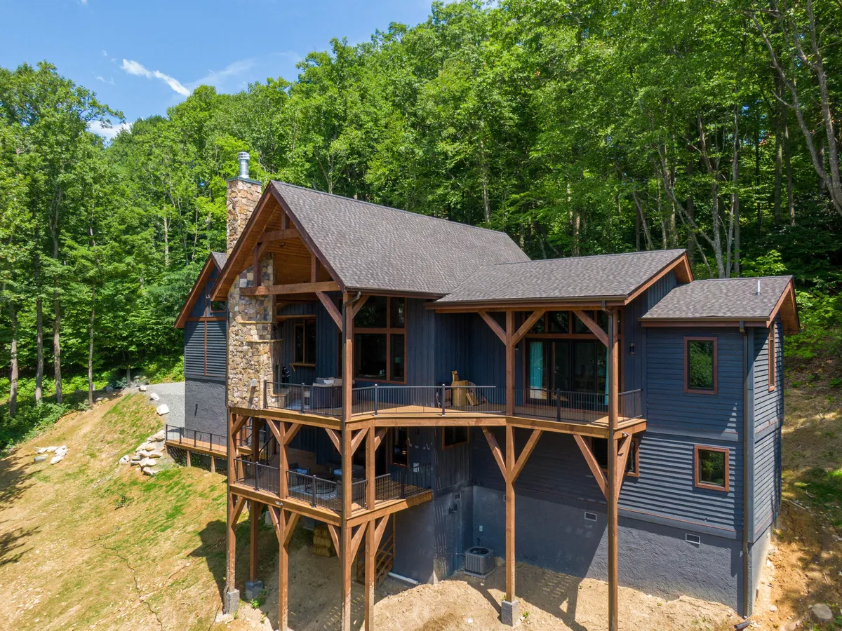 A wooden deck attached to the home overlooks the surrounding trees, with large windows and a stone chimney in view. The deck offers space for outdoor gatherings, relaxation, and family activities, connecting the home to the woods.