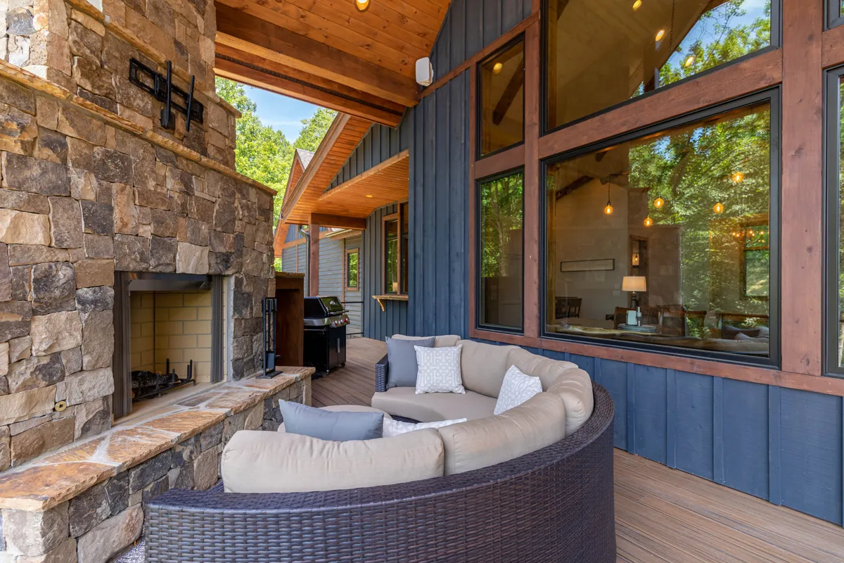 A cozy outdoor seating area arranged around a stone fireplace invites guests to relax beneath the open sky. Large windows of the home reflect the fading daylight, and the intimate setup is surrounded by mountain scenery, making it ideal for evening gatherings and conversations.