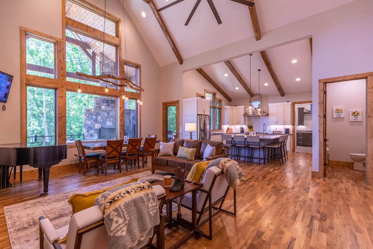 This image showcases a spacious, open-concept interior with large windows revealing a lush forest view. The central dining table, plush living area seating, and a grand piano create a warm and inviting space designed for gatherings, relaxation, and special moments with family or friends. The kitchen island and bar stools further highlight the space's entertainment potential.
