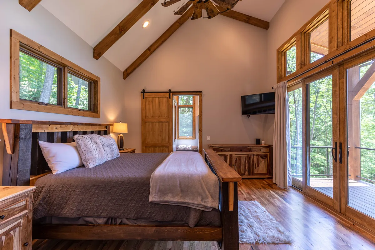 A tranquil bedroom featuring a plush bed with soft bedding, large windows framing views of nature, wooden furniture, and a ceiling fan overhead. Natural light fills the room, creating a serene and inviting atmosphere perfect for rest.
