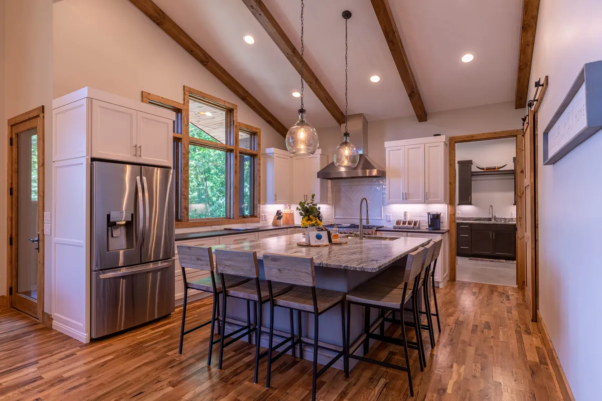 A spacious kitchen featuring a large island with granite counters, stainless steel appliances, wooden beams overhead, and expansive windows that flood the space with natural light. The setting is informal yet sophisticated, clearly designed for family gatherings and entertaining.