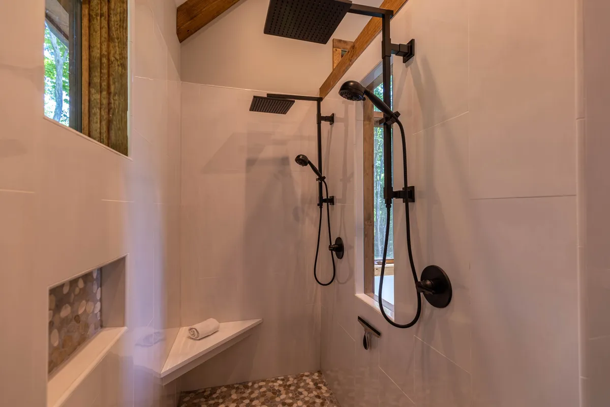 A luxurious bathroom featuring dual rainfall showerheads, a large window letting in abundant natural light, and textured pebble flooring. The spa-like atmosphere offers a refreshing, nature-immersed escape.