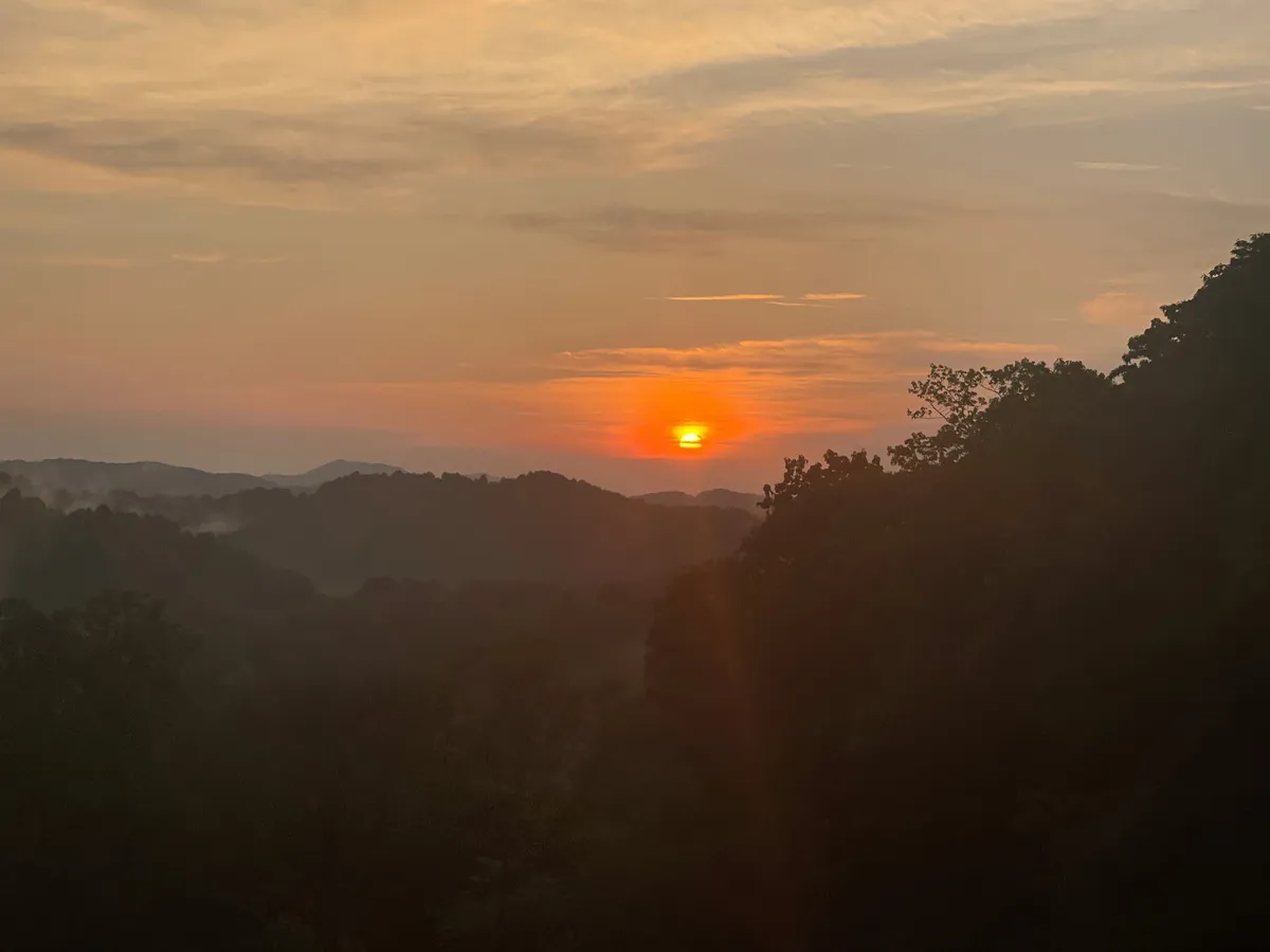 A spectacular sunset bathes the mountain range and clouds in soft, tranquil light, offering a panoramic view that invites relaxation. The expansive sky and distant peaks create a sense of peace and immersion in nature.