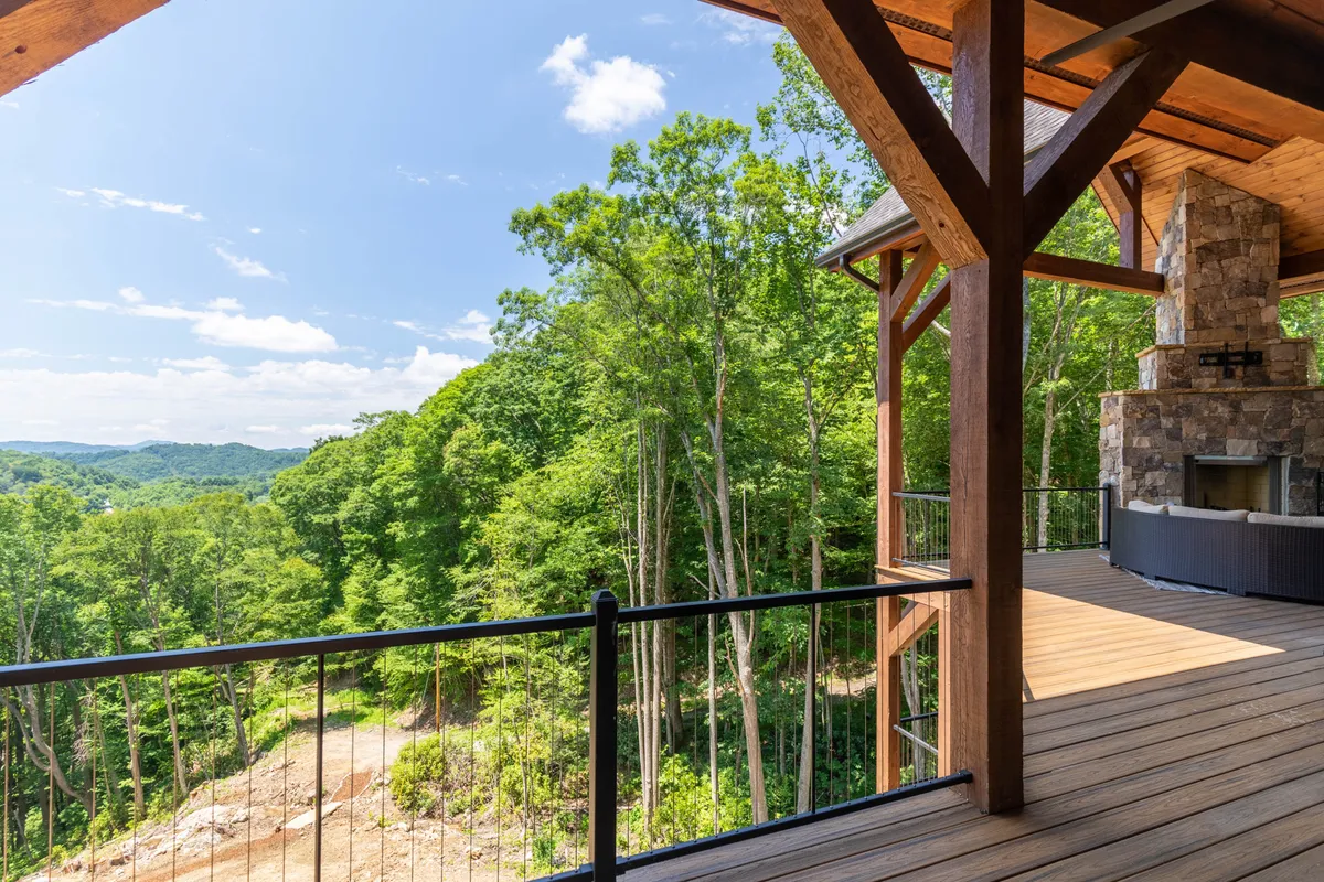 A covered wooden deck with a prominent stone fireplace, surrounded by lush greenery and offering distant views of rolling hills. Comfortable seating invites relaxation and conversation, highlighting the seamless transition between indoor and outdoor living.